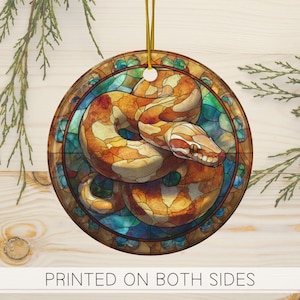 Op de afbeelding: Ronde ornament met een gedetailleerde illustratie van een slang in een glas-in-loodstijl. De slang is afgebeeld in tinten oranje, bruin en crème, met een gedetailleerde kop en een opgerold lichaam. Het ornament heeft een gouden touwtje om op te hangen en de tekst "PRINTED ON BOTH SIDES".