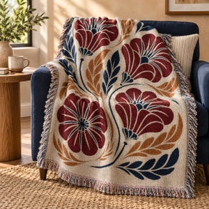 May include: A woven throw blanket featuring a cream-colored base with a large-scale floral pattern in deep red, navy blue, and golden yellow. The blanket has a textured appearance and is finished with decorative fringe along the edges.