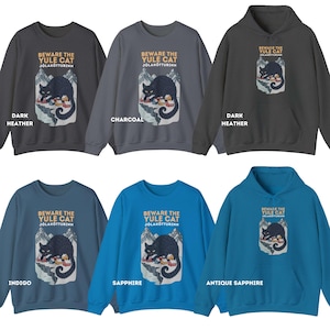 May include: A collection of six sweatshirts and hoodies in dark heather, charcoal, indigo, sapphire, and antique sapphire colors. Each garment features a graphic of a black cat over a snowy mountain village with the text BEWARE THE YULE CAT J&Oacute;LAK&Ouml;TTURINN.