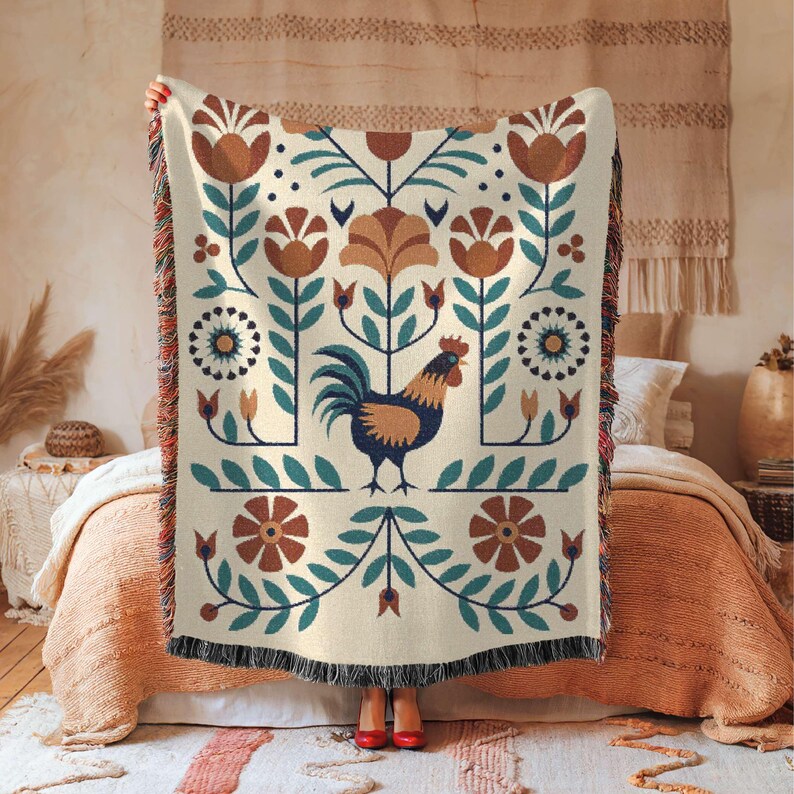 May include: Cream-coloured woven throw blanket featuring a rooster and floral design in shades of brown, teal, and blue. The blanket has a fringed edge and is held up in front of a bed.