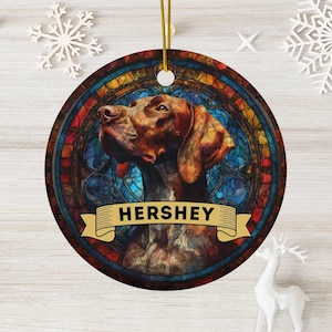 May include: Round Christmas ornament featuring a brown dog with the name "HERSHEY" on a banner. The dog is set against a stained-glass background with blue, red, and yellow hues. A gold string is attached for hanging.