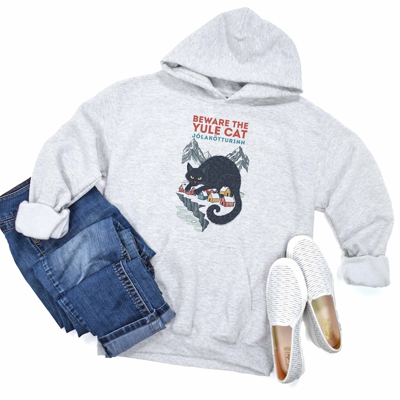 May include: A light gray hooded sweatshirt featuring a graphic of a black cat looming over a small village with mountains in the background. The text reads BEWARE THE YULE CAT and J&Oacute;LAK&Ouml;TTURINN. Also included are blue denim jeans and white slip-on shoes.