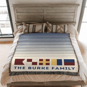 May include: A woven throw blanket with a nautical theme, featuring horizontal navy blue and white stripes. The bottom section displays nautical flags and the text "THE BURKE FAMILY" in navy blue. The blanket is fringed.