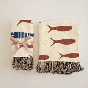 May include: Two woven throw blankets. One is rolled and tied with a tan ribbon. Both blankets have a cream background with brown fish designs and fringed edges. One blanket has a blue stripe.