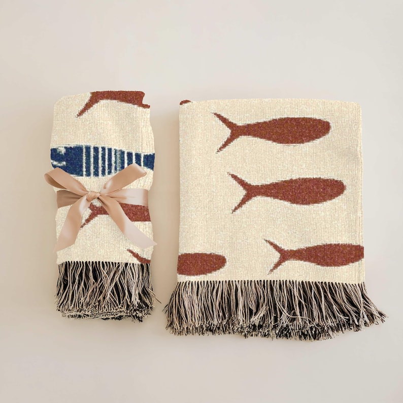 May include: Two woven throw blankets. One is rolled and tied with a tan ribbon. Both blankets have a cream background with brown fish designs and fringed edges. One blanket has a blue stripe.