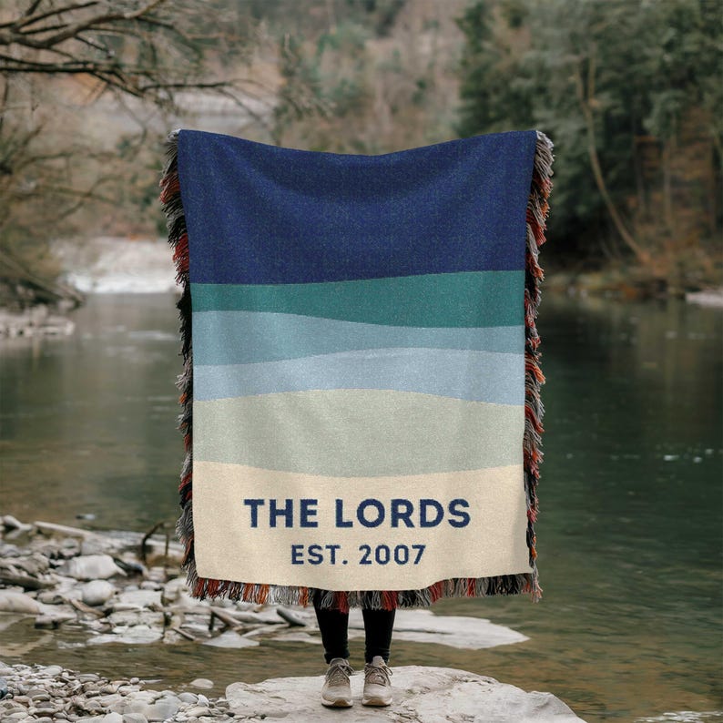 May include: A woven blanket with a gradient of blue and green horizontal stripes, with a cream-colored bottom section. The text "THE LORDS EST. 2007" is printed on the bottom. The blanket has fringed edges.