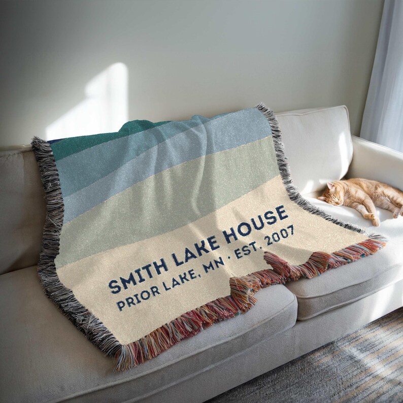 May include: A woven throw blanket with a gradient of blue, green, and beige stripes. The blanket has a fringed edge and text that reads "SMITH LAKE HOUSE PRIOR LAKE, MN EST. 2007". The blanket is draped over a white sofa.