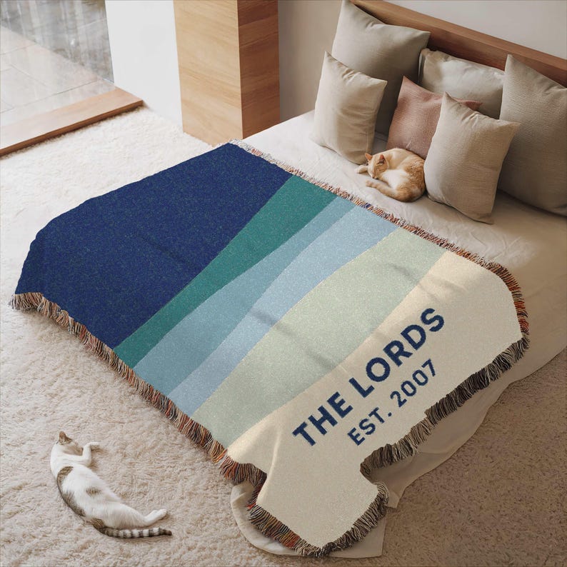 May include: A woven throw blanket with a gradient design in shades of blue and green. The blanket has a cream-colored section with the text "THE LORDS EST. 2007" in navy blue. The edges are fringed.