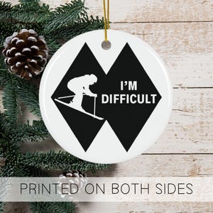 May include: White ceramic Christmas ornament with a black diamond shape and a skier silhouette. The text "I'm Difficult" is printed in white on the ornament.