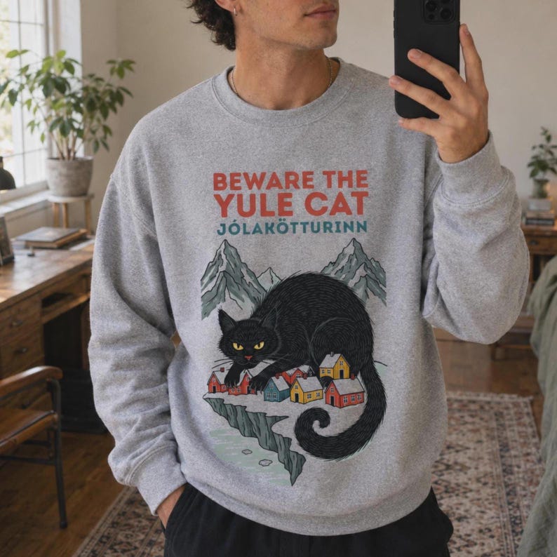 May include: A heather gray crewneck sweatshirt featuring a graphic print of a large black cat looming over a small village of colorful houses. Above the cat, the text reads BEWARE THE YULE CAT and J&Oacute;LAK&Ouml;TTURINN, with mountains in the background.