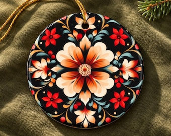 Puede incluir: Un adorno de cerámica redondo con fondo negro y un patrón floral simétrico en tonos naranja, crema, rojo y azul verdoso. El diseño incluye una flor central rodeada de flores más pequeñas y hojas en espiral, con un cordón dorado para colgar.