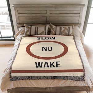 May include: A woven throw blanket with a white background and a red circle with the text "SLOW NO WAKE" in black.
