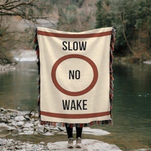 May include: A cream-colored woven throw blanket with a red stripe border. The blanket features a red circle with the words "SLOW NO WAKE" in black text.