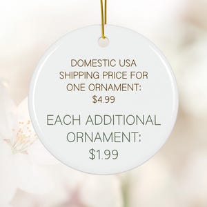 May include: A white, round ceramic ornament with a gold string for hanging. The ornament has text in brown, including "DOMESTIC USA" and "EACH ADDITIONAL ORNAMENT". The background is a soft, blurred floral pattern.