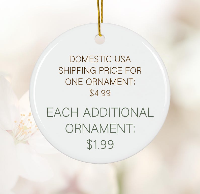 May include: A white, round ceramic ornament with a gold string for hanging. The ornament has text in brown, including "DOMESTIC USA" and "EACH ADDITIONAL ORNAMENT". The background is a soft, blurred floral pattern.