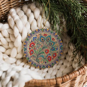 May include: Round Christmas ornament with a floral design in red, blue, and green. The ornament has a gold string for hanging and is set against a white knitted blanket and a wicker basket.