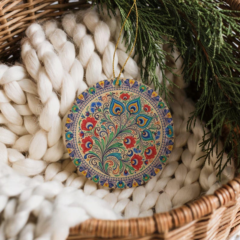 May include: Round Christmas ornament with a floral design in red, blue, and green. The ornament has a gold string for hanging and is set against a white knitted blanket and a wicker basket.