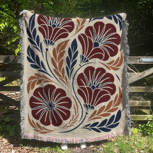 May include: A woven throw blanket featuring a beige base with a large-scale floral pattern in deep red, navy blue, and burnt orange. The design includes stylized flowers and elongated leaves, finished with a decorative fringed edge on all sides.