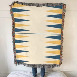 Könnte beinhalten: Eine gewebte Wolldecke mit einem geometrischen Muster aus gelben und blauen Dreiecken. Die Decke hat an allen Seiten Fransen.