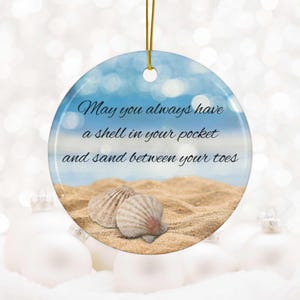 May include: A round ceramic ornament with a blue and white beach scene and seashells. The ornament has the text "May you always have a shell in your pocket and sand between your toes."