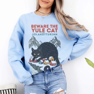 May include: A light blue crewneck sweatshirt featuring a graphic print of a black cat crouching over a small snowy village with mountains in the background. The text reads BEWARE THE YULE CAT J&Oacute;LAK&Ouml;TTURINN in red and teal lettering on the front.