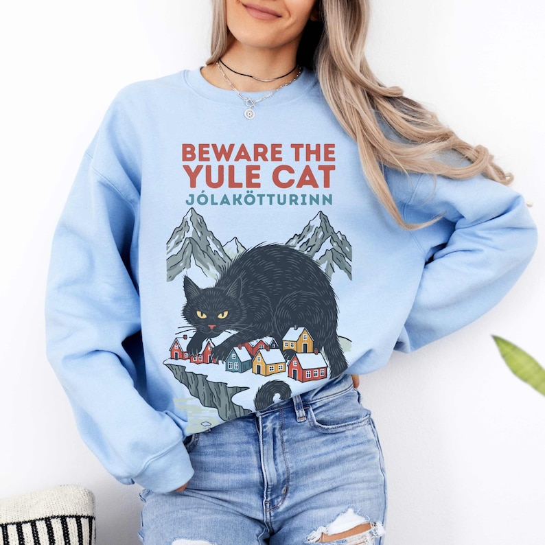 May include: A light blue crewneck sweatshirt featuring a graphic print of a black cat crouching over a small snowy village with mountains in the background. The text reads BEWARE THE YULE CAT J&Oacute;LAK&Ouml;TTURINN in red and teal lettering on the front.