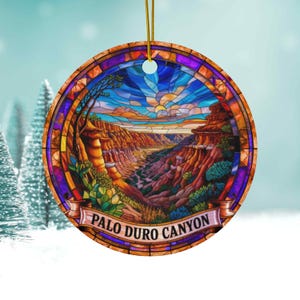 May include: Round ceramic ornament featuring a stained-glass style depiction of Palo Duro Canyon. The design includes a canyon landscape with a river, blue sky, and the text "PALO DURO CANYON" on a banner. A gold string is attached for hanging.