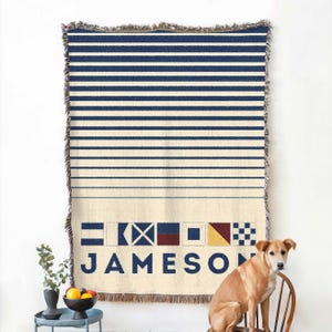 May include: Cream-colored throw blanket with navy blue horizontal stripes and fringed edges. The bottom features nautical signal flags and the name "JAMESON" in navy blue. A dog sits on a wooden chair next to the blanket.