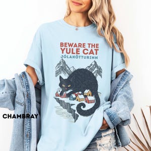 May include: A light blue t-shirt featuring a graphic of a large black cat looming over a small village of colorful houses nestled on a cliffside with mountains in the background. The text reads BEWARE THE YULE CAT J&Oacute;LAK&Ouml;TTURINN in red and blue.