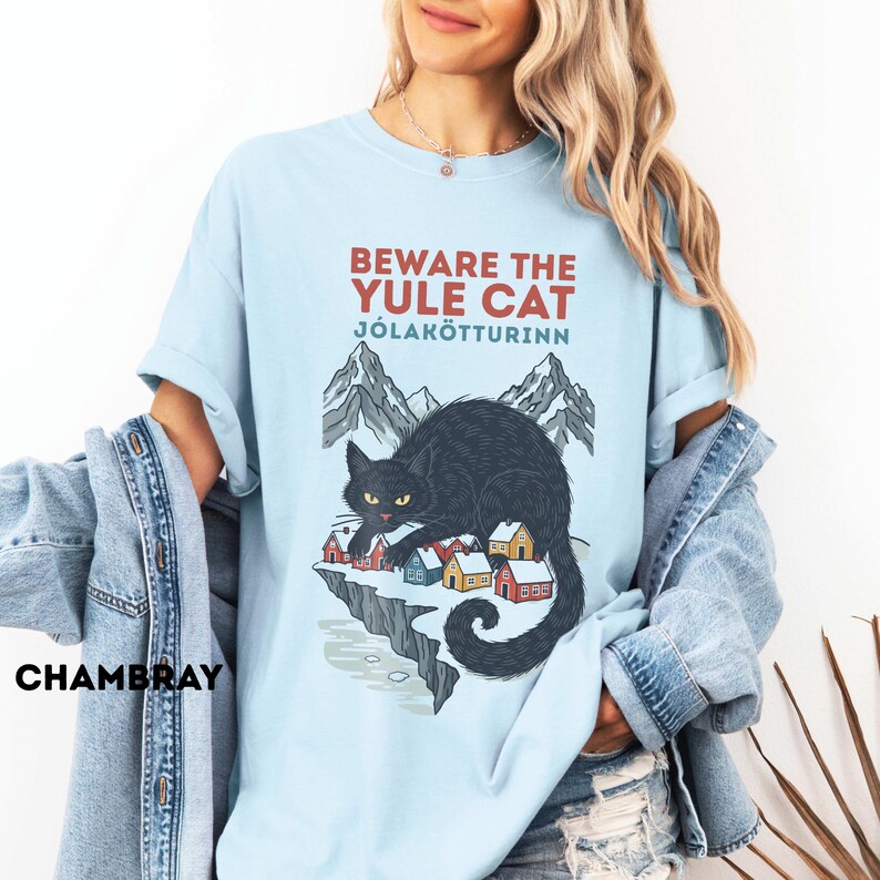 May include: A light blue t-shirt featuring a graphic of a large black cat looming over a small village of colorful houses nestled on a cliffside with mountains in the background. The text reads BEWARE THE YULE CAT J&Oacute;LAK&Ouml;TTURINN in red and blue.