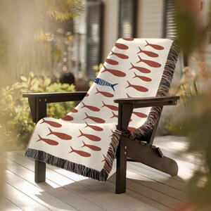 May include: A cream-colored throw blanket draped over a dark wooden Adirondack chair. The blanket features a repeating pattern of rust-colored fish, with one blue fish. The blanket has a fringed edge.