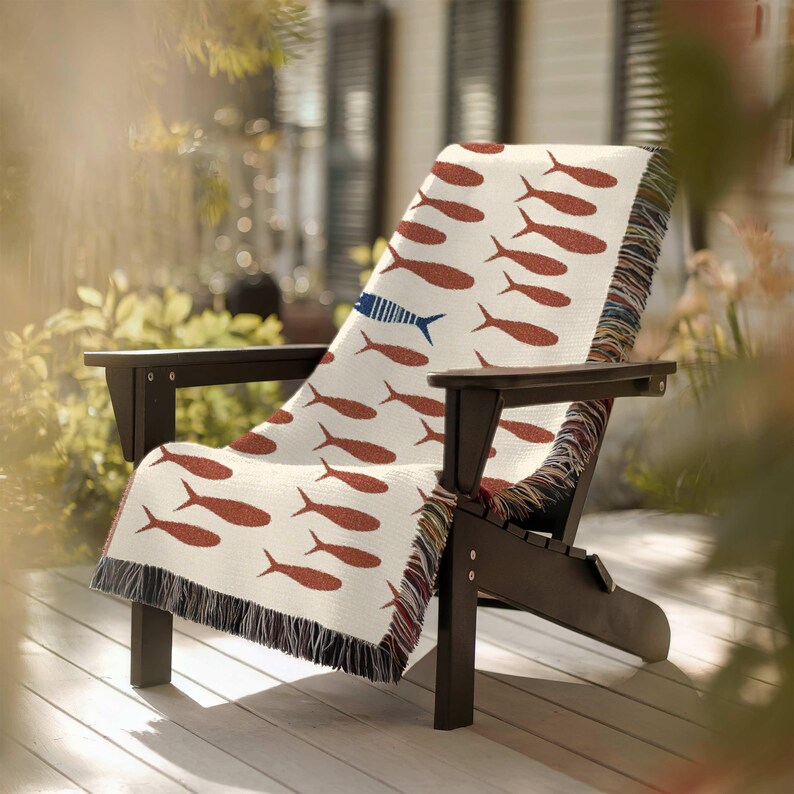 May include: A cream-colored throw blanket draped over a dark wooden Adirondack chair. The blanket features a repeating pattern of rust-colored fish, with one blue fish. The blanket has a fringed edge.