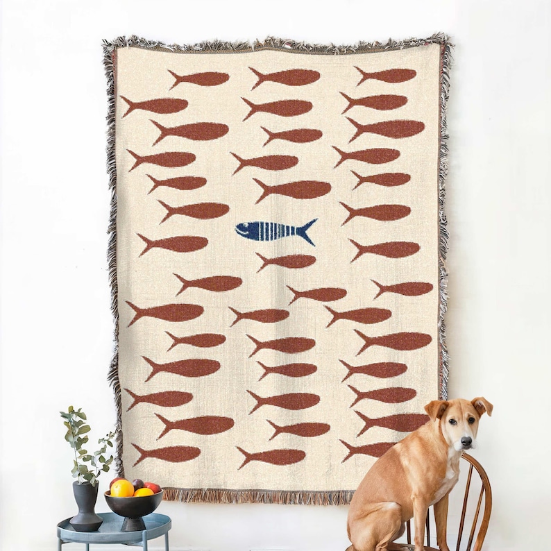 May include: Cream-colored woven throw blanket with a repeating pattern of brown fish. One blue fish with vertical stripes stands out. The blanket has a fringed edge and is displayed on a wall.
