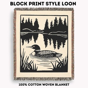 May include: Cream-colored woven blanket with a black block print design. The design features a loon swimming in a lake with a forest backdrop. The text "BLOCK PRINT STYLE LOON" and "100% COTTON WOVEN BLANKET" are at the top and bottom.