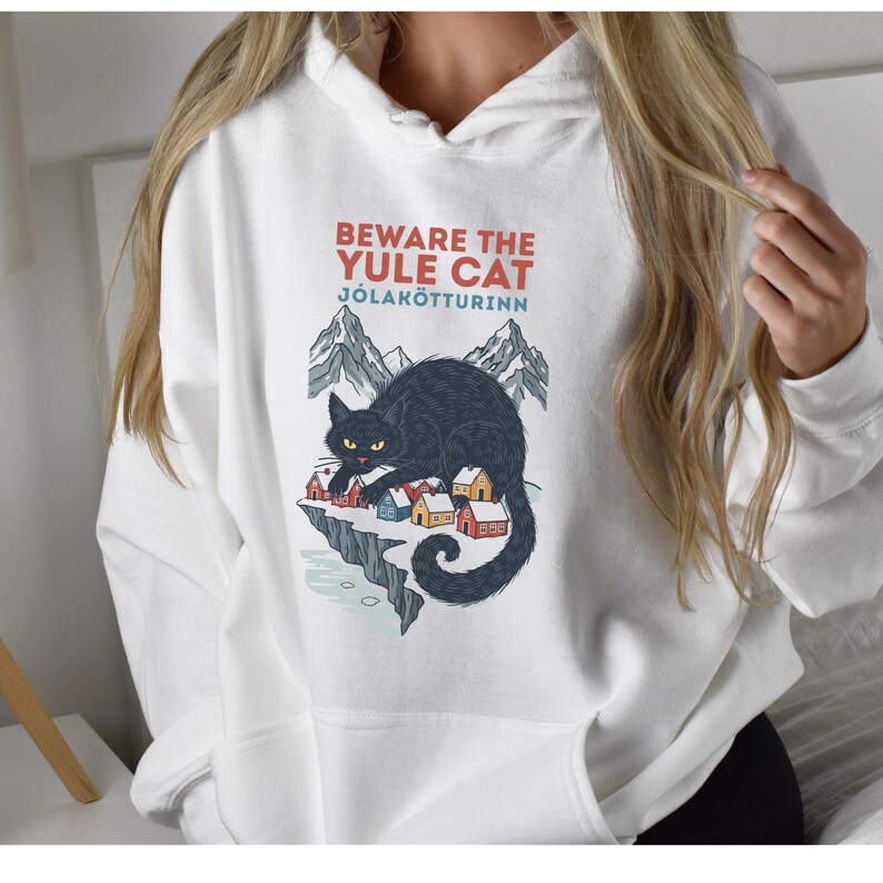 May include: A white hooded sweatshirt featuring a graphic print of a large black cat looming over a small village with snowy mountains in the background. The text reads BEWARE THE YULE CAT J&Oacute;LAK&Ouml;TTURINN in red and blue lettering above the illustration.