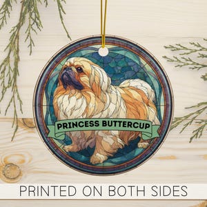 May include: Round wooden ornament featuring a Pekingese dog illustration. The dog is depicted in shades of brown and cream, set against a stained-glass style background. The text "PRINCESS BUTTERCUP" is on a green banner. The bottom text reads "PRINTED ON BOTH SIDES".