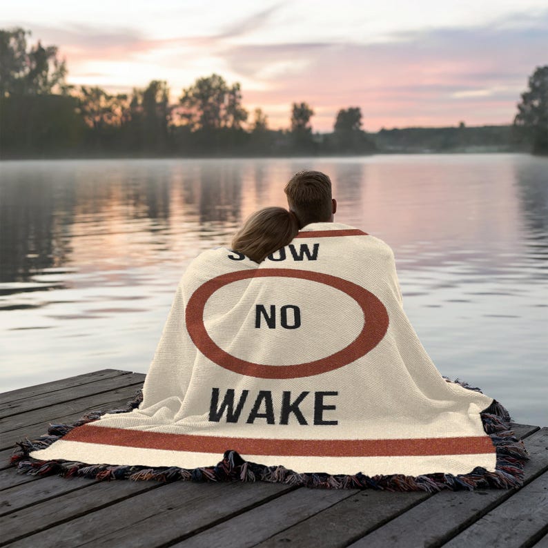 May include: A couple sits on a wooden dock wrapped in a cream-colored throw blanket with a red stripe and the words "Slow No Wake" printed on it.