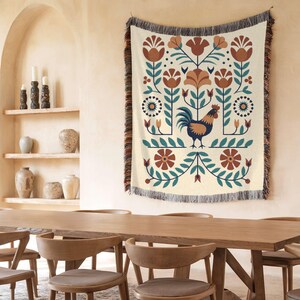 May include: A cream-coloured woven throw blanket with a folk art design featuring a rooster, flowers, and foliage in shades of brown, teal, and blue. The blanket has a fringed edge and is hanging on a wall.