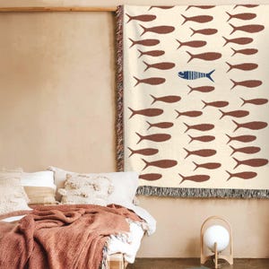 May include: A cream-colored throw blanket with a repeating pattern of brown fish. One blue fish stands out among the brown ones. The blanket has a fringed edge and is displayed on a bed with pillows and a terracotta-colored blanket.