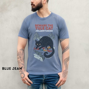 May include: A dusty blue short-sleeve t-shirt featuring a graphic of a large black cat looming over a small village with mountains in the background. The text reads BEWARE THE YULE CAT J&Oacute;LAK&Ouml;TTURINN in red and white lettering on the chest.
