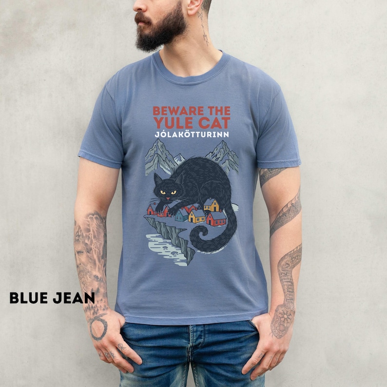 May include: A dusty blue short-sleeve t-shirt featuring a graphic of a large black cat looming over a small village with mountains in the background. The text reads BEWARE THE YULE CAT J&Oacute;LAK&Ouml;TTURINN in red and white lettering on the chest.