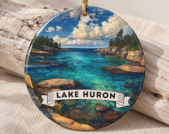 May include: A round ceramic ornament featuring a vibrant illustration of Lake Huron with clear turquoise water, rocky shorelines, and a blue sky with fluffy white clouds. A white banner across the bottom displays the text LAKE HURON in black.
