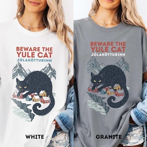 May include: A white and a granite gray t-shirt, each featuring a graphic of a black cat perched on a snowy cliff above a small village. The text reads BEWARE THE YULE CAT and J&Oacute;LAK&Ouml;TTURINN in red and blue lettering above the mountain illustration.