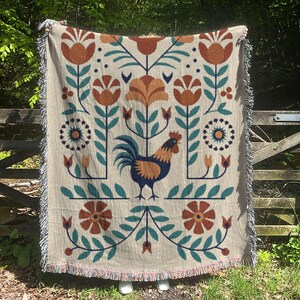 May include: Beige woven throw blanket with a repeating floral and rooster design. The design features orange, teal, and navy blue elements. The blanket has a fringed edge and is displayed outdoors.