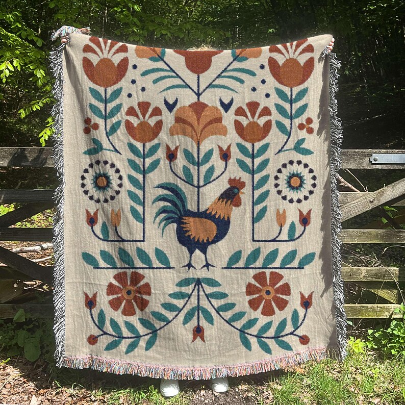 May include: Beige woven throw blanket with a repeating floral and rooster design. The design features orange, teal, and navy blue elements. The blanket has a fringed edge and is displayed outdoors.