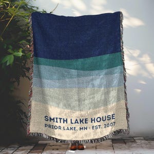 May include: A woven blanket with horizontal stripes in shades of blue, green, and beige. The blanket has a fringed edge and the text "SMITH LAKE HOUSE PRIOR LAKE, MN EST. 2007" in navy blue.