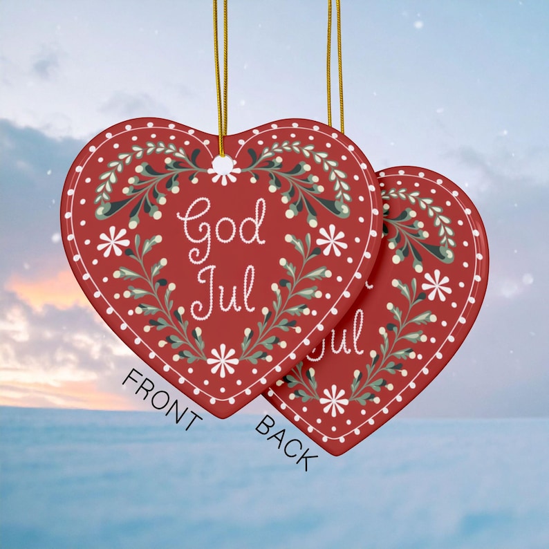 May include: Two heart-shaped Christmas ornaments. The ornaments are red with white floral designs and the words "God Jul" in white script. Gold string is attached for hanging. The word "FRONT" and "BACK" are printed on the ornaments.