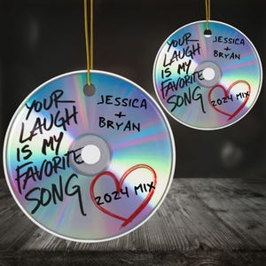 以下が含まれることがあります： 「Your laugh is my favorite song」と「2024 Mix」の文字が書かれた銀色のCDオーナメント2つ。文字は黒のマーカーで書かれており、各オーナメントには赤いハートが描かれている。