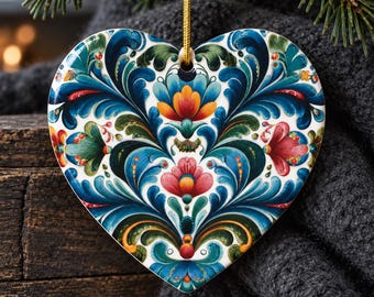 May include: A heart-shaped ceramic ornament featuring a symmetrical folk art pattern with intricate blue, teal, green, orange, and red floral motifs. The ornament hangs from a thin gold cord and displays a glossy finish with detailed scrollwork.