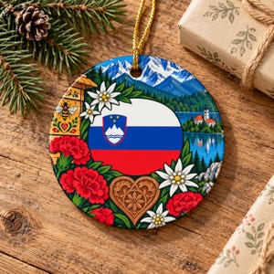 Op de afbeelding: Een rond keramisch ornament met de Sloveense vlag in het midden, omringd door illustraties van rode anjers, witte edelweissbloemen, een houten bijenkorf met een bij, een hartvormig houtsnijwerk en een bergmeerlandschap.